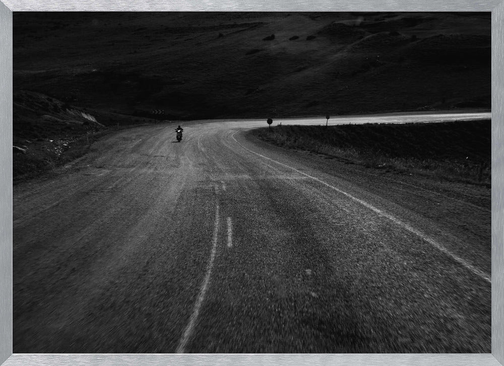 A moody, black and white photograph capturing a lone motorcyclist from behind as they navigate a curving road through a dark, hilly terrain, framed in silver. Decor