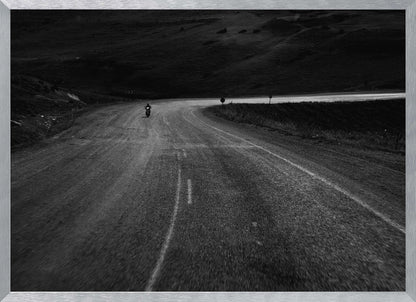 A moody, black and white photograph capturing a lone motorcyclist from behind as they navigate a curving road through a dark, hilly terrain, framed in silver. Decor