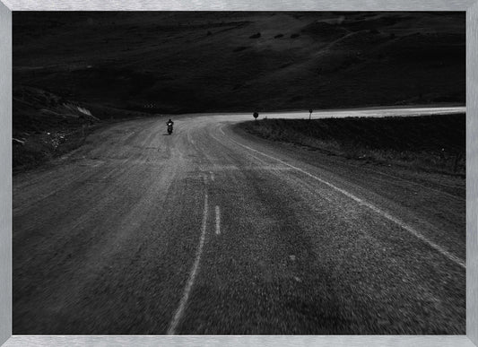 A moody, black and white photograph capturing a lone motorcyclist from behind as they navigate a curving road through a dark, hilly terrain, framed in silver. Decor