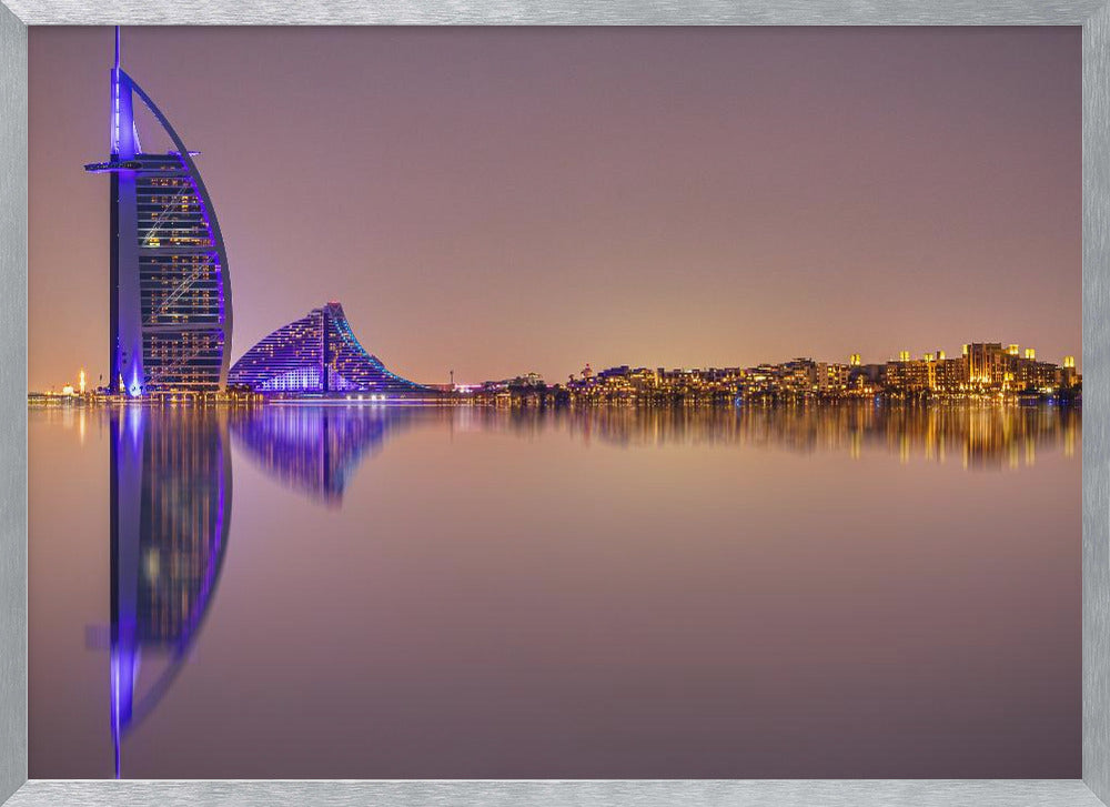 A framed panoramic night photograph of the Dubai skyline featuring the Burj Al Arab hotel lit up in brilliant purple. The city lights and architecture are perfectly reflected in the calm water in the foreground, all under a hazy, dusky sky. Poster