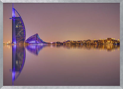 A framed panoramic night photograph of the Dubai skyline featuring the Burj Al Arab hotel lit up in brilliant purple. The city lights and architecture are perfectly reflected in the calm water in the foreground, all under a hazy, dusky sky. Poster