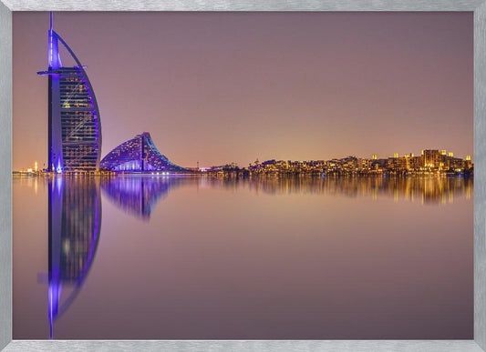 A framed panoramic night photograph of the Dubai skyline featuring the Burj Al Arab hotel lit up in brilliant purple. The city lights and architecture are perfectly reflected in the calm water in the foreground, all under a hazy, dusky sky. Poster