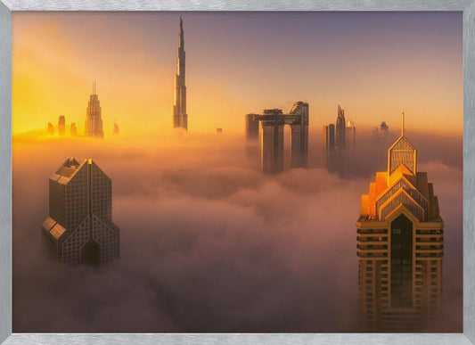 An aerial photograph of the Dubai skyline shrouded in a thick blanket of fog at sunrise. The warm golden light illuminates the tops of several skyscrapers, including the Burj Khalifa, which pierce through the clouds creating a dreamlike and majestic scene. Print