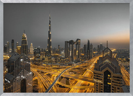 A framed, high-angle, long-exposure photograph of the Dubai skyline at dusk. The towering Burj Khalifa stands out against a grey sky, while the city's skyscrapers are illuminated. In the foreground, a complex network of highways glows with the yellow light trails of traffic, creating intricate patterns. Poster