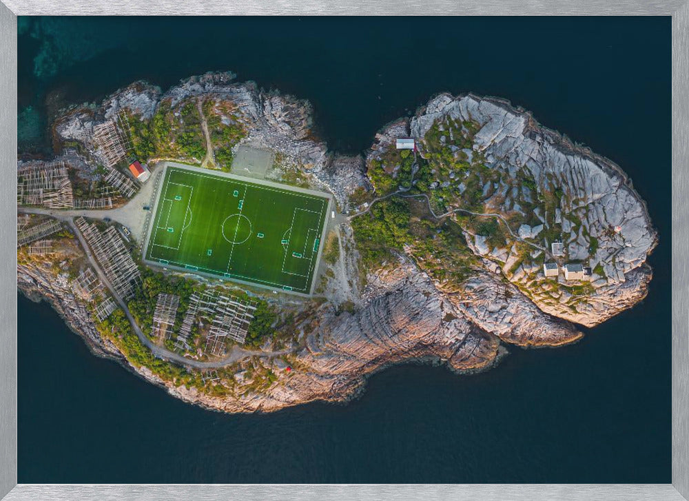 An aerial view of a vibrant green soccer field situated on a small, rocky island surrounded by dark blue water. The photograph, taken from directly above, captures the unique and remote location of the sports facility, with small buildings and wooden structures dotting the island's terrain. The entire scene is enclosed within a silver frame. Print