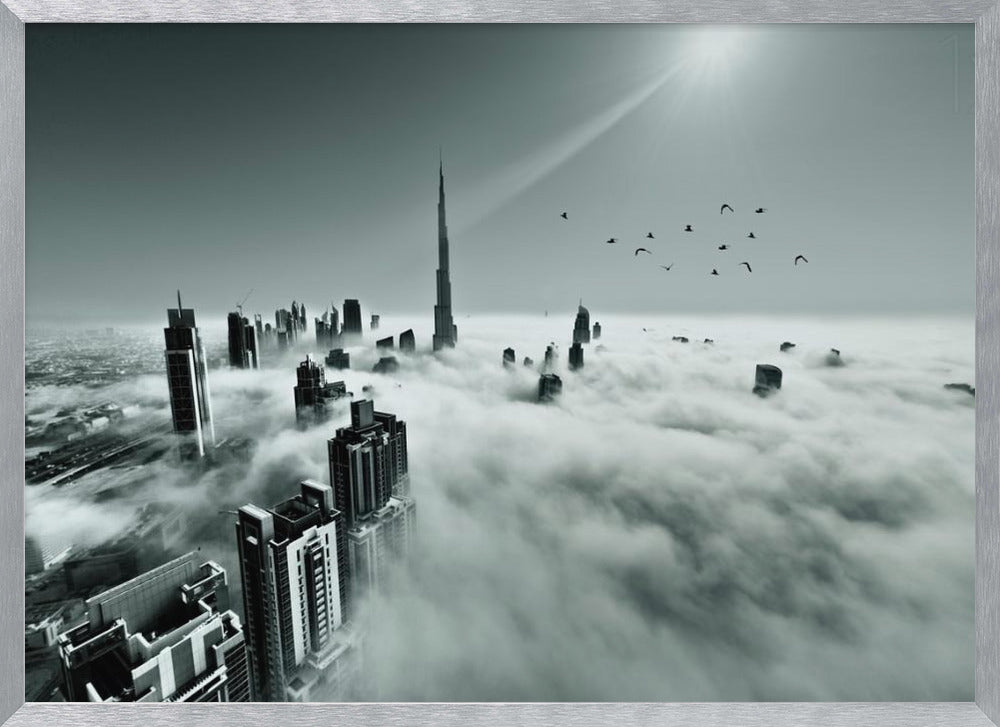 A framed, black and white aerial photograph of the Dubai skyline piercing through a thick layer of clouds. The Burj Khalifa stands prominently in the center, with other skyscrapers emerging from the fog. A flock of birds flies in the sky as a bright sunbeam shines down from the upper right corner. Wall Art