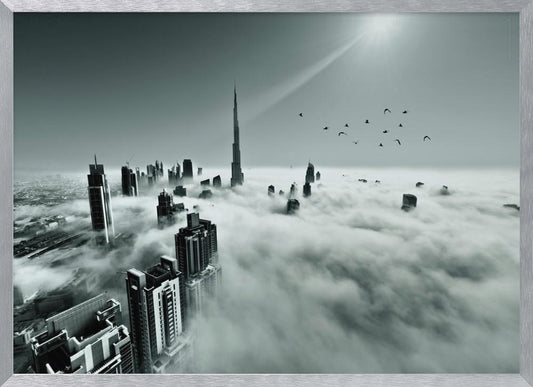 A framed, black and white aerial photograph of the Dubai skyline piercing through a thick layer of clouds. The Burj Khalifa stands prominently in the center, with other skyscrapers emerging from the fog. A flock of birds flies in the sky as a bright sunbeam shines down from the upper right corner. Wall Art