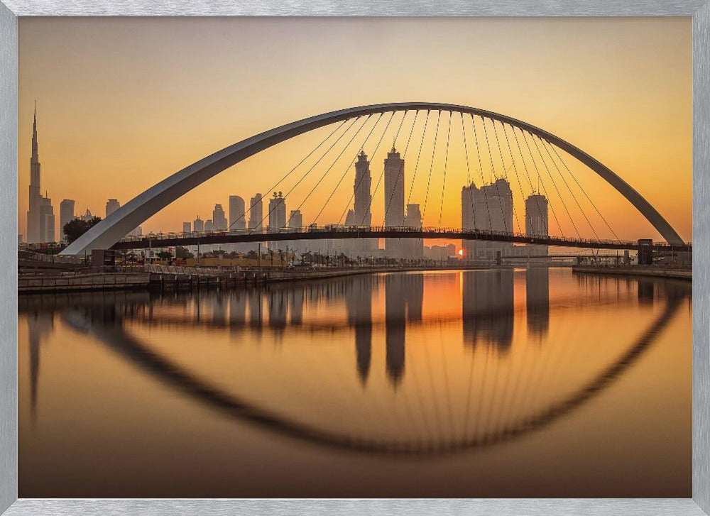 A panoramic photograph of the Dubai skyline at sunrise, viewed from across the water. A large, modern arched bridge spans the canal in the foreground, and its reflection creates a perfect oval shape in the calm water. In the background, skyscrapers, including the Burj Khalifa, are silhouetted against a glowing orange and yellow sky. The entire scene is presented within a silver picture frame. Wall Art