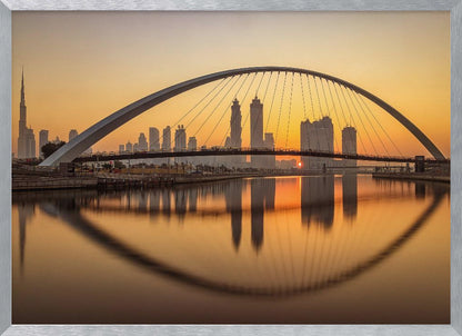 A panoramic photograph of the Dubai skyline at sunrise, viewed from across the water. A large, modern arched bridge spans the canal in the foreground, and its reflection creates a perfect oval shape in the calm water. In the background, skyscrapers, including the Burj Khalifa, are silhouetted against a glowing orange and yellow sky. The entire scene is presented within a silver picture frame. Wall Art