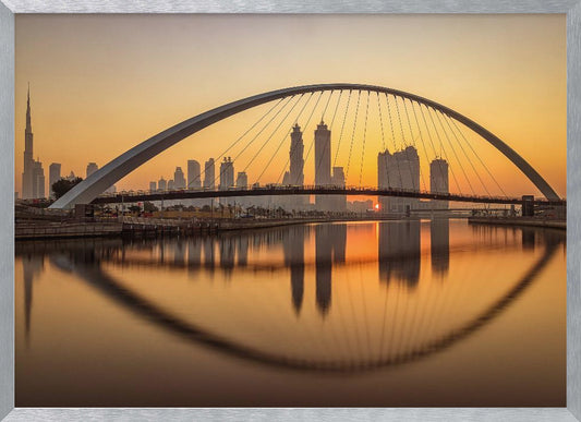 A panoramic photograph of the Dubai skyline at sunrise, viewed from across the water. A large, modern arched bridge spans the canal in the foreground, and its reflection creates a perfect oval shape in the calm water. In the background, skyscrapers, including the Burj Khalifa, are silhouetted against a glowing orange and yellow sky. The entire scene is presented within a silver picture frame. Wall Art
