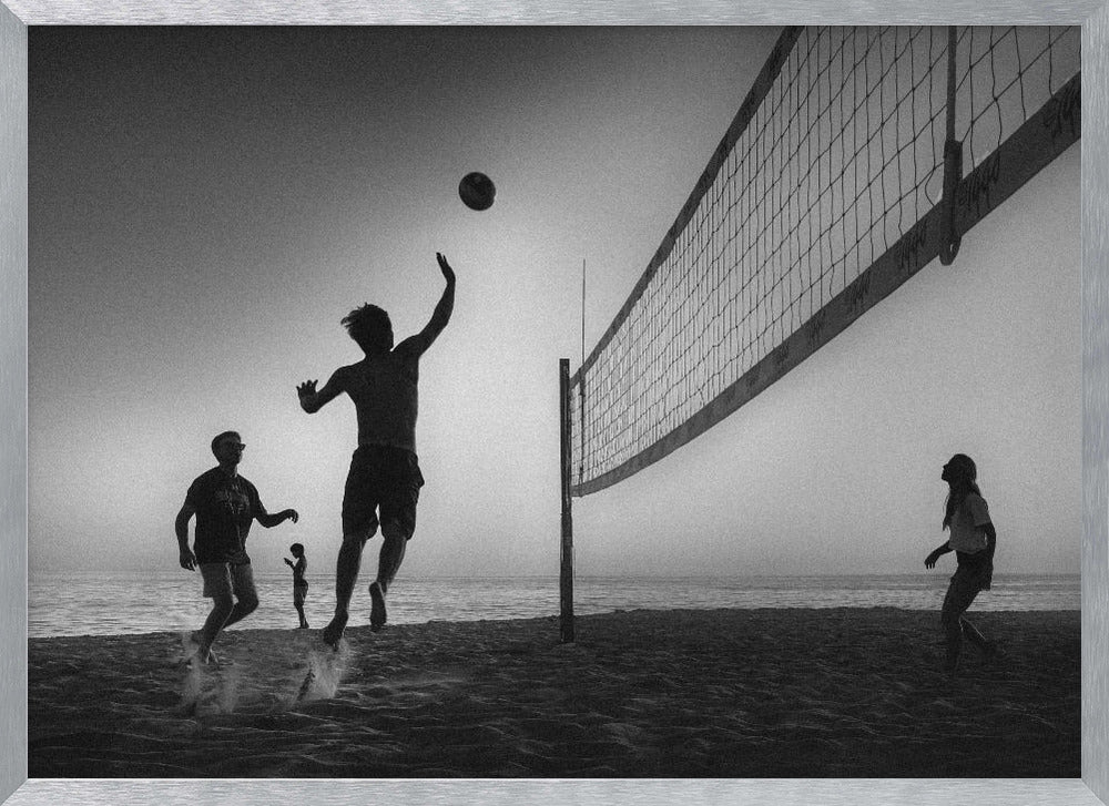 A black and white action shot of people playing volleyball on a beach, presented in a silver frame. The scene is silhouetted against a light sky, likely during sunrise or sunset. One player is captured mid-jump, arm raised to hit the ball over the net. Other players are visible on the sand, with the ocean in the background. Decor