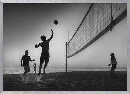 A black and white action shot of people playing volleyball on a beach, presented in a silver frame. The scene is silhouetted against a light sky, likely during sunrise or sunset. One player is captured mid-jump, arm raised to hit the ball over the net. Other players are visible on the sand, with the ocean in the background. Decor