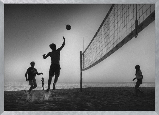 A black and white action shot of people playing volleyball on a beach, presented in a silver frame. The scene is silhouetted against a light sky, likely during sunrise or sunset. One player is captured mid-jump, arm raised to hit the ball over the net. Other players are visible on the sand, with the ocean in the background. Decor