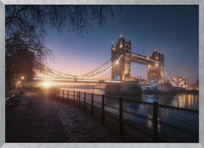 A scenic photograph of London's Tower Bridge at sunrise, its lights glowing warmly against a deep blue and orange sky. In the foreground, a cobblestone path follows the River Thames, leading the eye towards the iconic bridge and the rising sun. Poster