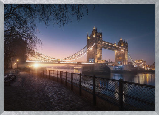 A scenic photograph of London's Tower Bridge at sunrise, its lights glowing warmly against a deep blue and orange sky. In the foreground, a cobblestone path follows the River Thames, leading the eye towards the iconic bridge and the rising sun. Poster
