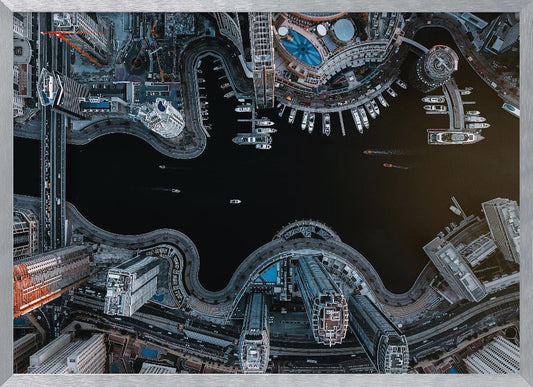 A dramatic top-down aerial photograph of a sprawling modern city at night. A dark waterway curves through the center, surrounded by illuminated skyscrapers, busy roads, and a marina packed with boats, all within a silver picture frame. Artwork