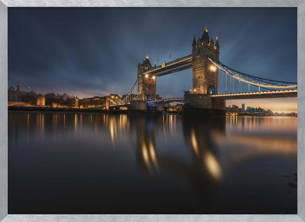 A long-exposure photograph of London's Tower Bridge at dusk, brilliantly illuminated with golden lights that cast shimmering reflections on the calm River Thames. The sky is a deep, dark blue, and the city skyline glows in the background, all enclosed within a silver frame. Print