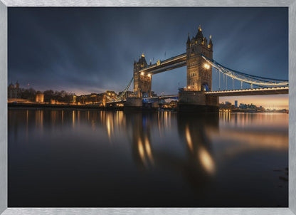 A long-exposure photograph of London's Tower Bridge at dusk, brilliantly illuminated with golden lights that cast shimmering reflections on the calm River Thames. The sky is a deep, dark blue, and the city skyline glows in the background, all enclosed within a silver frame. Print
