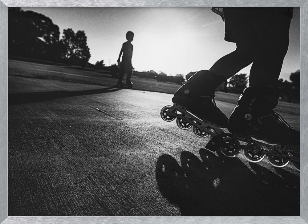 A low-angle, black and white photograph of two people rollerblading on a paved path at sunset. The foreground is a close-up of one person's rollerblades and their long shadow, while the second person is silhouetted in the distance against the bright sun. Artwork