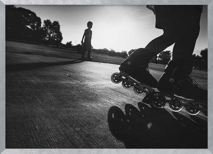 A low-angle, black and white photograph of two people rollerblading on a paved path at sunset. The foreground is a close-up of one person's rollerblades and their long shadow, while the second person is silhouetted in the distance against the bright sun. Artwork