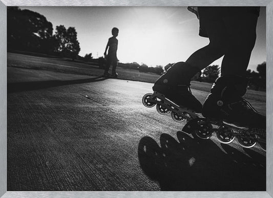 A low-angle, black and white photograph of two people rollerblading on a paved path at sunset. The foreground is a close-up of one person's rollerblades and their long shadow, while the second person is silhouetted in the distance against the bright sun. Artwork