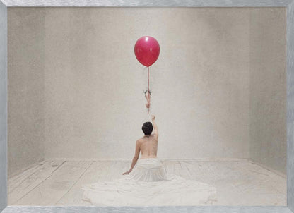 A surreal fine art photograph of a person with a bare back and a long white skirt, sitting on the floor of a minimalist grey room. They are reaching up towards a small figure hanging from the string of a large, bright red balloon that floats towards the ceiling. The image is enclosed in a silver frame. Artwork