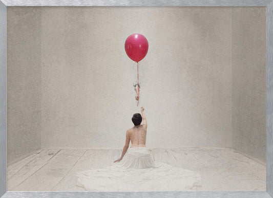 A surreal fine art photograph of a person with a bare back and a long white skirt, sitting on the floor of a minimalist grey room. They are reaching up towards a small figure hanging from the string of a large, bright red balloon that floats towards the ceiling. The image is enclosed in a silver frame. Artwork