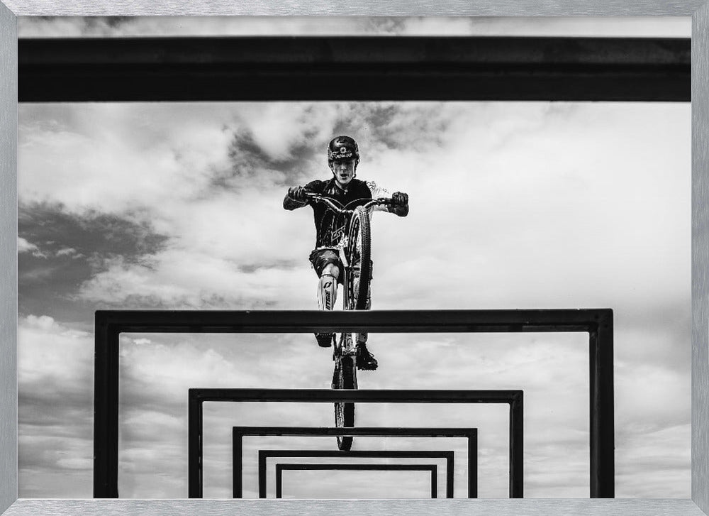 A striking black and white photograph taken from a low angle, showing a cyclist in full gear balancing on a mountain bike. The cyclist is positioned centrally above a series of black rectangular frames that create a receding, tunnel-like visual effect against a cloudy sky. The entire image is presented within a silver picture frame. Artwork