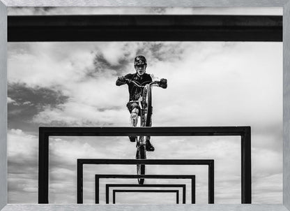 A striking black and white photograph taken from a low angle, showing a cyclist in full gear balancing on a mountain bike. The cyclist is positioned centrally above a series of black rectangular frames that create a receding, tunnel-like visual effect against a cloudy sky. The entire image is presented within a silver picture frame. Artwork