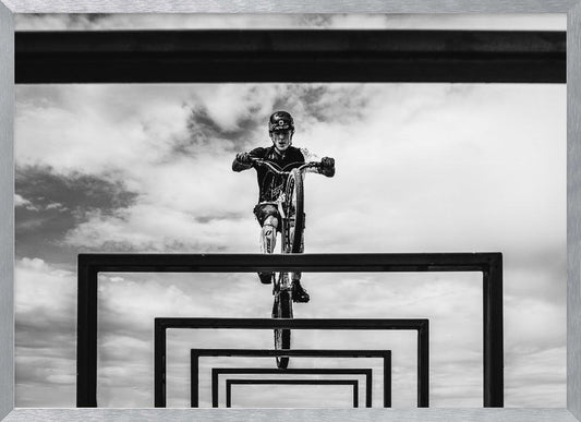 A striking black and white photograph taken from a low angle, showing a cyclist in full gear balancing on a mountain bike. The cyclist is positioned centrally above a series of black rectangular frames that create a receding, tunnel-like visual effect against a cloudy sky. The entire image is presented within a silver picture frame. Artwork