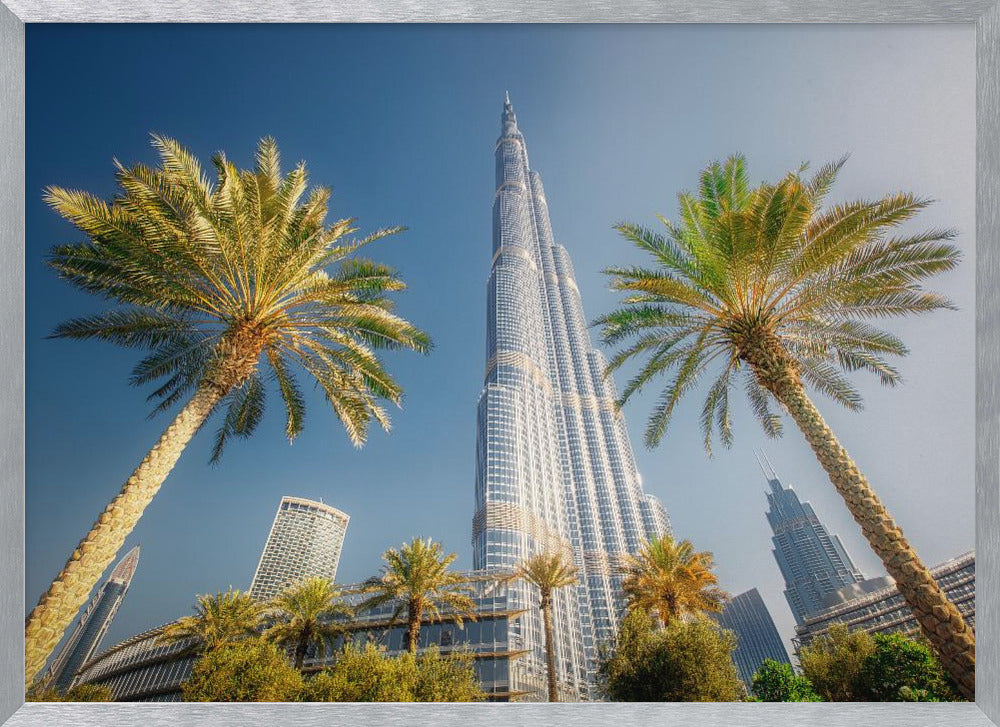 A low-angle photograph looking up at the towering Burj Khalifa skyscraper in Dubai, framed by two large palm trees against a clear, vibrant blue sky. Wall Art
