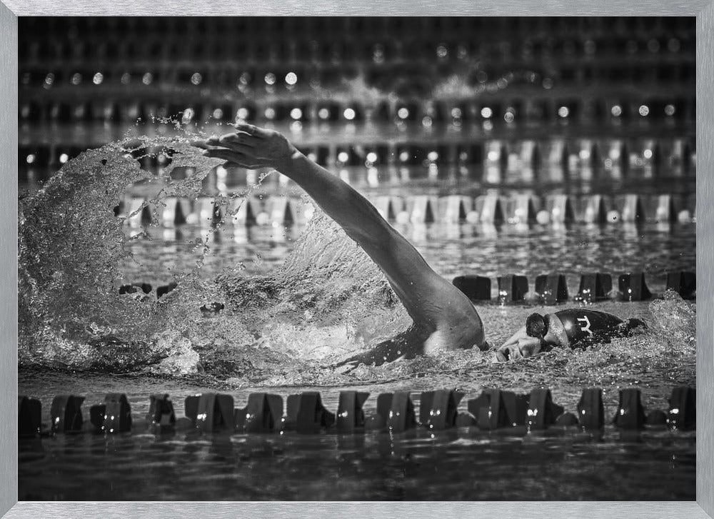 A dramatic black and white action photograph of a competitive swimmer mid-stroke, captured within a silver frame. The swimmer's arm is extended forward, creating a powerful splash of water. The focus is on the motion and intensity of the sport, with the pool's lane lines blurred in the background. Wall Art