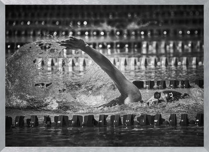 A dramatic black and white action photograph of a competitive swimmer mid-stroke, captured within a silver frame. The swimmer's arm is extended forward, creating a powerful splash of water. The focus is on the motion and intensity of the sport, with the pool's lane lines blurred in the background. Wall Art