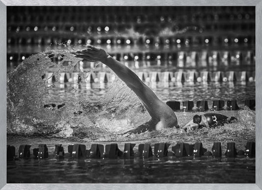 A dramatic black and white action photograph of a competitive swimmer mid-stroke, captured within a silver frame. The swimmer's arm is extended forward, creating a powerful splash of water. The focus is on the motion and intensity of the sport, with the pool's lane lines blurred in the background. Wall Art