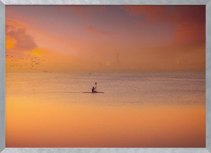 A serene photograph of a lone kayaker silhouetted against a vast expanse of calm water during a hazy, golden sunset. The sky and water are bathed in warm hues of orange, pink, and purple. A flock of birds flies in the distance, and the faint outline of a city skyline is visible on the horizon. Wall Art