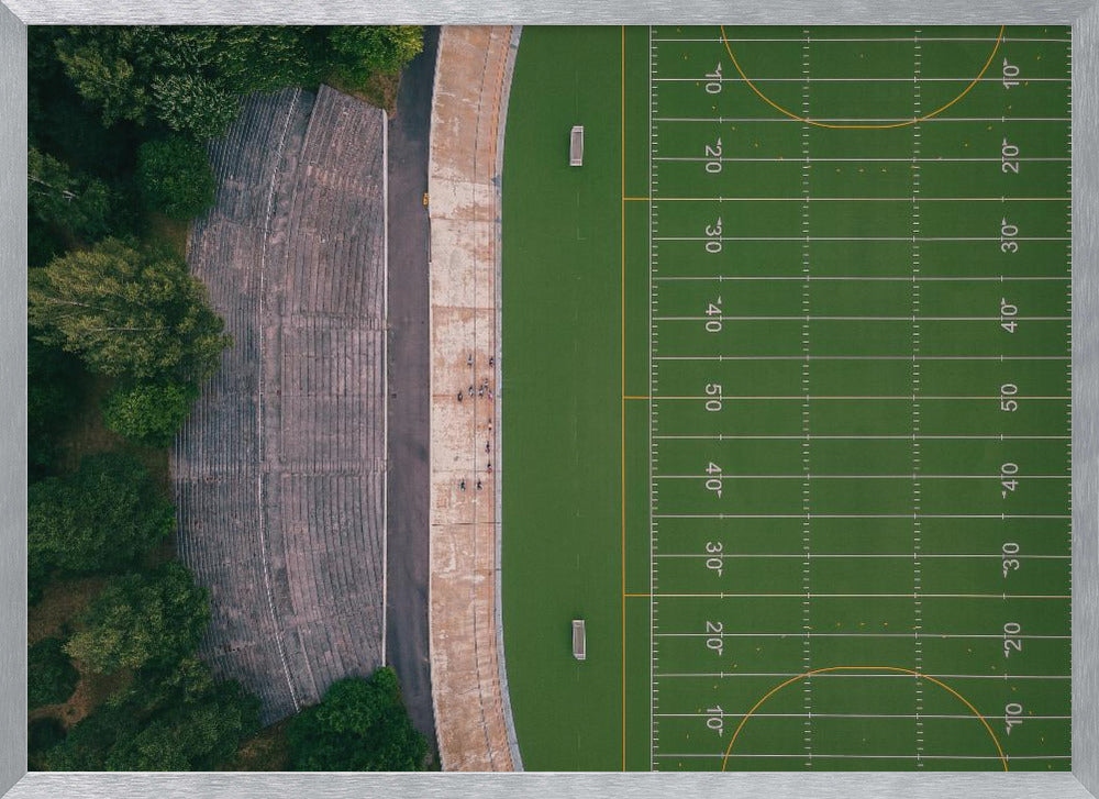 An aerial top-down photograph of an American football field beside empty, weathered concrete stadium bleachers. The vibrant green field is marked with white yard lines, and the gray stands are nestled amongst lush green trees. The entire scene is enclosed in a silver frame. Artwork