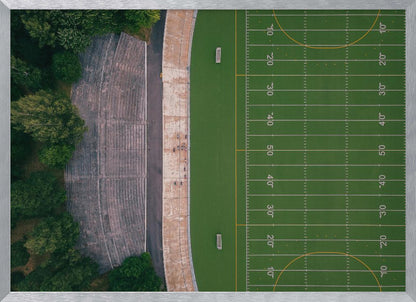 An aerial top-down photograph of an American football field beside empty, weathered concrete stadium bleachers. The vibrant green field is marked with white yard lines, and the gray stands are nestled amongst lush green trees. The entire scene is enclosed in a silver frame. Artwork