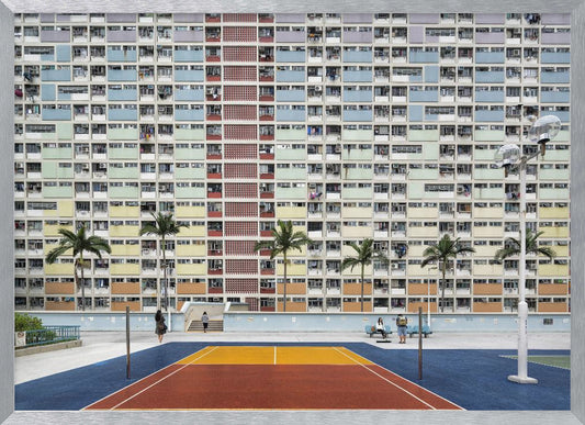A wide, symmetrical photograph of the colorful Choi Hung Estate apartment building in Hong Kong, featuring a facade with a repeating pattern of pastel blue, green, yellow, and orange blocks. In front of the building is a vibrant basketball court painted in bold sections of blue, red, and yellow, with several palm trees lining the courtyard. Print