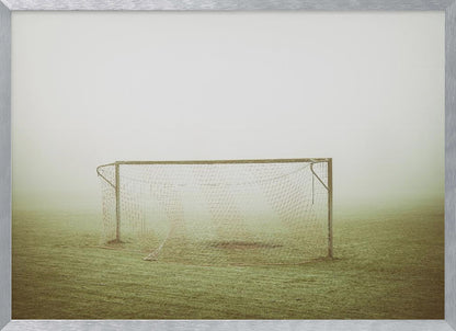 A framed photograph of an empty soccer goal with a net, standing alone on a green grass field enveloped in thick, white fog, creating a minimalist and atmospheric scene. Poster