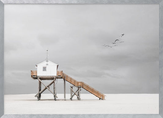 A minimalist, desaturated photograph of a small white cabin on tall wooden stilts on a vast, empty beach. A long wooden staircase leads up to the cabin. The sky is a flat, overcast grey, and a flock of dark birds is visible flying in the distance. Print