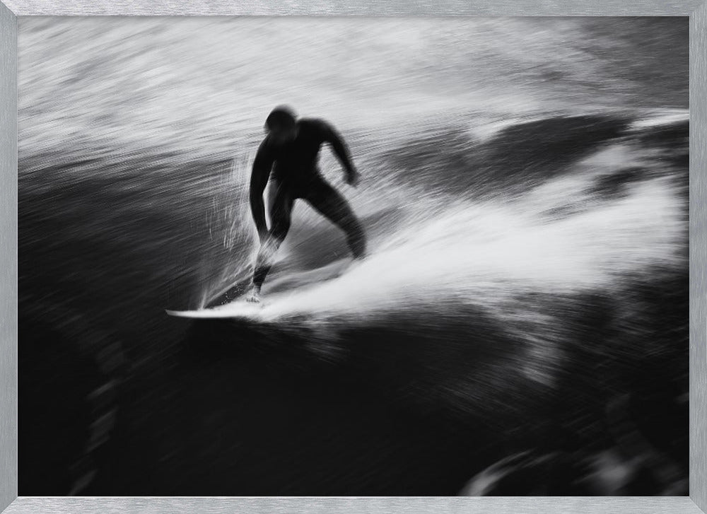 A dynamic black and white photograph of a surfer in a wetsuit carving through a wave. The image uses motion blur to emphasize the speed and energy of the water, creating a powerful and artistic effect. The artwork is presented in a simple silver frame. Print