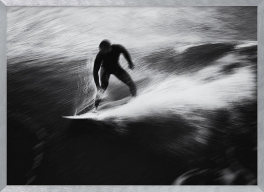 A dynamic black and white photograph of a surfer in a wetsuit carving through a wave. The image uses motion blur to emphasize the speed and energy of the water, creating a powerful and artistic effect. The artwork is presented in a simple silver frame. Print