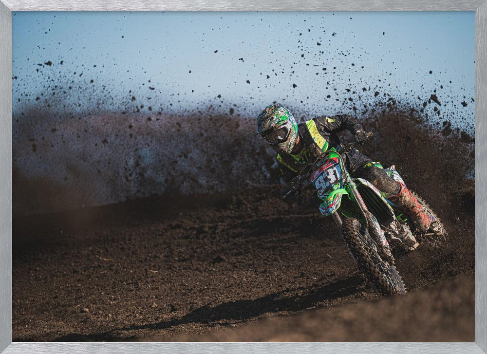 A motocross rider in green and black gear leans hard into a turn on a dirt track, kicking up a large spray of mud against a clear blue sky. Decor
