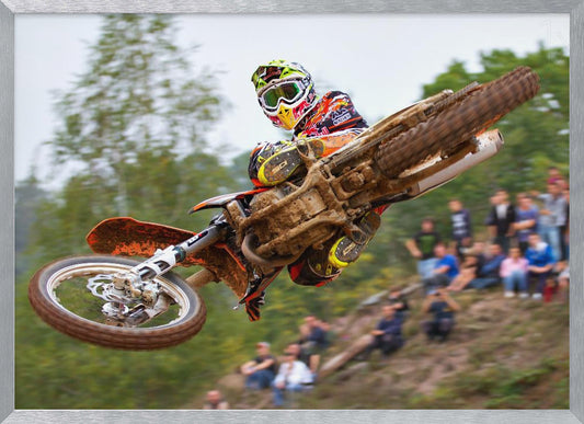 A dynamic action shot of a motocross racer in mid-air, leaning into a turn with their muddy orange dirt bike. The rider wears a colorful helmet and racing gear, with a blurred background of green trees and spectators watching from a dirt hill. The photo is enclosed in a silver frame. Decor