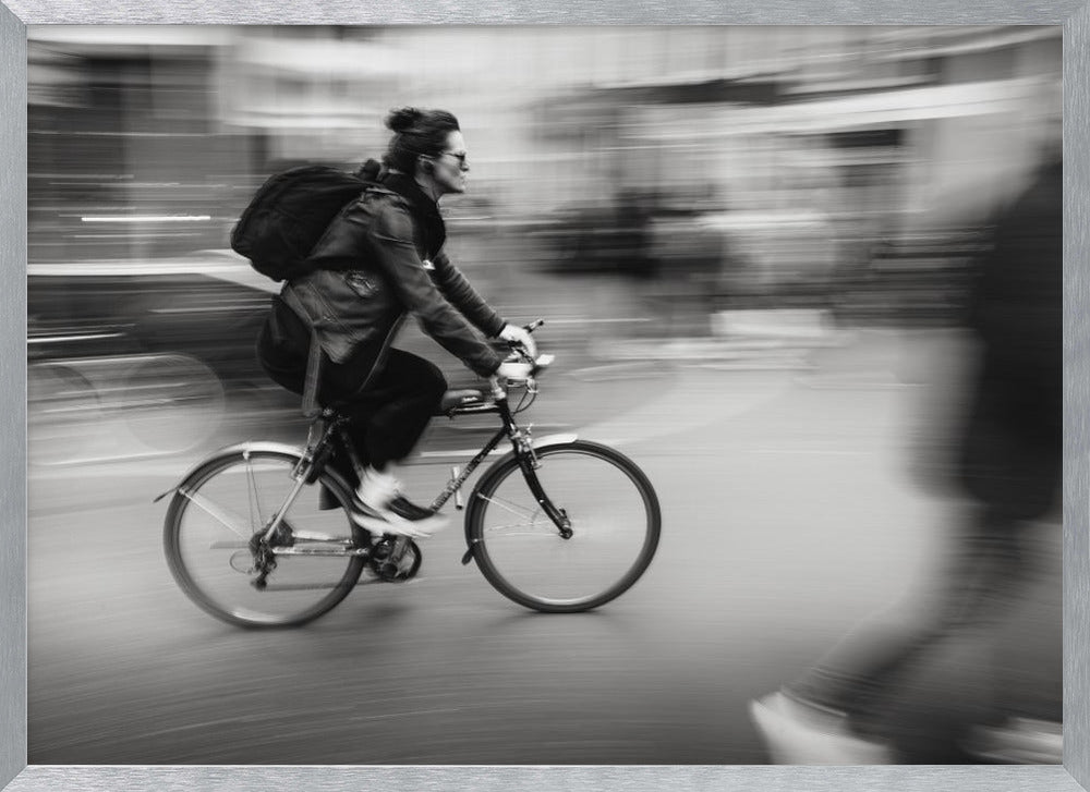 A dynamic black and white photograph capturing a person in profile as they cycle swiftly through a city. The background is a motion blur of urban scenery, highlighting the cyclist's speed. The individual wears a leather jacket, sunglasses, a backpack, and has their hair tied up. The artwork is enclosed in a silver frame. Wall Art
