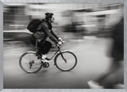 A dynamic black and white photograph capturing a person in profile as they cycle swiftly through a city. The background is a motion blur of urban scenery, highlighting the cyclist's speed. The individual wears a leather jacket, sunglasses, a backpack, and has their hair tied up. The artwork is enclosed in a silver frame. Wall Art