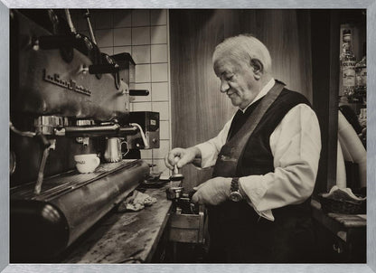 A sepia-toned, framed photograph of an elderly barista with white hair, wearing a vest, as he skillfully prepares an espresso. He is focused on his task, tamping the coffee grounds with a gentle smile, standing next to a large, vintage-style espresso machine in a cozy cafe setting. Print
