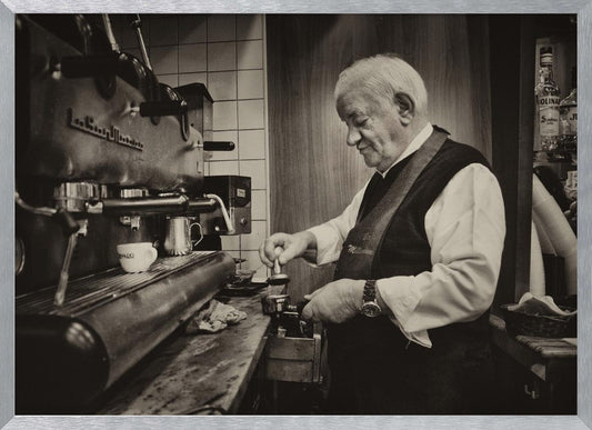 A sepia-toned, framed photograph of an elderly barista with white hair, wearing a vest, as he skillfully prepares an espresso. He is focused on his task, tamping the coffee grounds with a gentle smile, standing next to a large, vintage-style espresso machine in a cozy cafe setting. Print