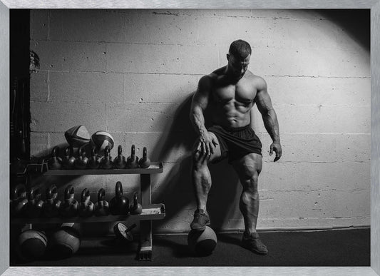A dramatic black and white photograph of a shirtless, muscular bodybuilder posing in a gym. He leans against a cinder block wall with one foot propped on a medicine ball, his chiseled physique and defined muscles highlighted by intense lighting. Racks of kettlebells are visible in the background. Wall Art