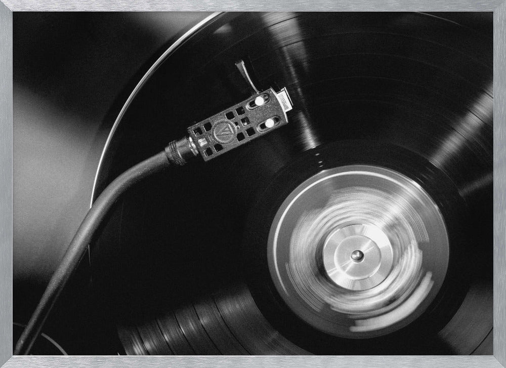 A framed, black and white, close-up photograph of a record player in action. The turntable is spinning a vinyl record, creating a motion blur on the center label. The tonearm and stylus are positioned over the grooved surface of the record. Poster